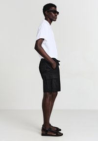 Uomo con occhiali da sole neri, camicia bianca a maniche corte, pantaloni cargo neri e sandali neri, in piedi di profilo contro uno sfondo semplice.