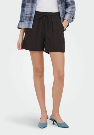 Kvinde iført sorte shorts med snøre, blå ternet skjorte over hvid top og blå, lavhælede spidse sko, stående med den ene hånd i lommen.