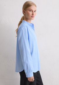 Blondynka z kucykiem ubraną w luźną, jasnoniebieską koszulę na guziki i czarne spodnie, stoi bokiem na tle białej ściany.