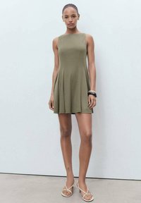 O rochie fără mâneci, de culoare verde măslinie, realizată dintr-un material neted, având o fustă evazată care se extinde până la mijlocul coapsei, cu cusături curate.