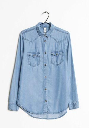 Jasnoniebieska jeansowa koszula z długim rękawem, zapinana na guziki z przodu, z dwoma kieszeniami na piersi z klapkami zapinanymi na guziki i zaostrzonym kołnierzem, wisząca na czarnym wieszaku.