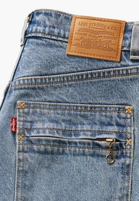 Levi's farkut vaaleansinisinä, joissa on ruskea nahkalaatta, vetoketjullinen tasku, kultaiset tikkauslangat ja punainen lappu sivulla.