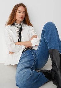 Camicia bianca a maniche lunghe, jeans a gamba larga blu denim, stivali in pelle nera e sciarpa a fantasia bianco e nero. Modello seduto con postura rilassata.