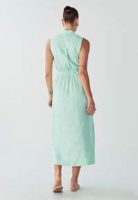 Mėtų žalios spalvos be rankovių suknelė su apykakle, elastinga juosmena ir ilgu, plazdančiu siluetu. Lygus audinys su subtiliu blizgesiu.