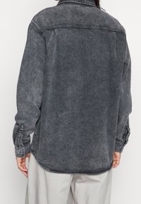 Osoba v opraném černém džínovém saku s knoflíkovými manžetami, zobrazená zezadu, kombinující s světle šedými kalhotami.