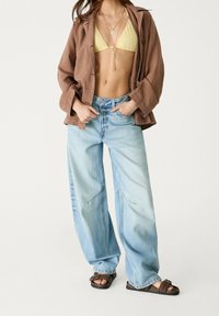 Veste en lin marron, haut de bikini jaune côtelé et jean à jambes larges bleu clair avec des accents déchirés, associ é à des sandales marron.