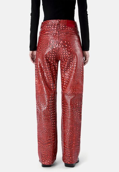 Personne portant un pantalon large brillant à motif crocodile rouge et un haut à manches longues noir, vue de dos sur un fond uni.