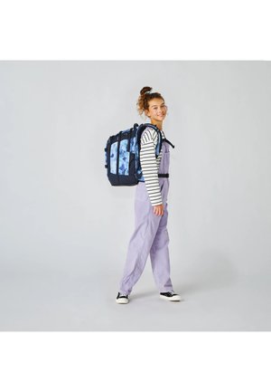 Junge Person, die lila Overalls und ein gestreiftes Hemd trägt, trägt einen großen blauen Rucksack mit Muster und steht vor einem schlichten grauen Hintergrund.