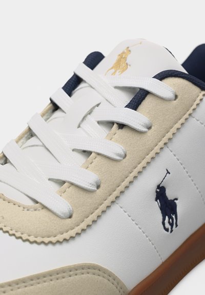 Άσπρο αθλητικό παπούτσι με μπεζ γούνινη επένδυση, λευκά κορδόνια, σκούρο μπλε και χρυσό λογότυπο Polo, και καστανή σόλα, εμφανιζόμενο σε κοντινή λήψη.