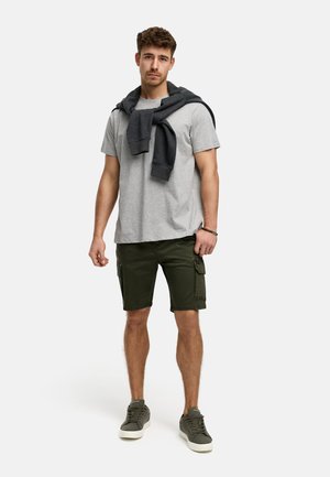 Szary bawełniany t-shirt, ciemnoszary sweter narzucony na ramiona, zielone szorty cargo oraz szare sneakersy z białymi podeszwami.