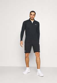 Svart långärmad träningsskjorta med dragkedja fram, kombinerad med svarta shorts. Båda plaggen har en logotyp. Vita sneakers kompletterar outfiten.