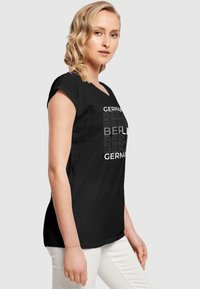 Sort T-shirt med v-halsdesign, der har hvid tekst, som skriver "TYSKLAND" og "BERLIN" i forskellige størrelser, parret med lyse bukser.