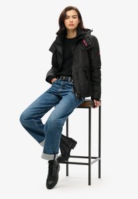 Czarna kurtka puchowa z zamkiem i detalami naszywkami, w połączeniu z niebieskimi dżinsami i czarnymi sznurowanymi botkami. Model siedzi na stołku barowym.
