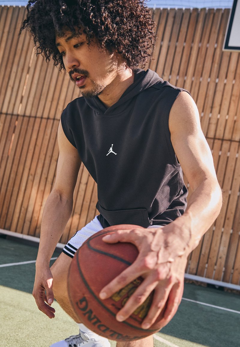 Sort ærmeløs hoodie med en frontlomme og hvidt logo, parret med hvide shorts med sorte striber, og en basketball på en bane.
