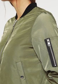 Zbliżenie oliwkowej kurtki bombowca z czarnym prążkowanym kołnierzem, przednim zamkiem błyskawicznym, kieszenią na rękawie z zamkiem oraz kieszenią boczną na zatrzask.