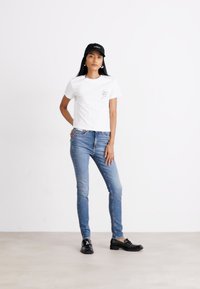 Fehér, rövidített póló grafikai mintával, világoskék skinny farmerral és fekete mokaszinnal párosítva. A modellen fekete sapka van.