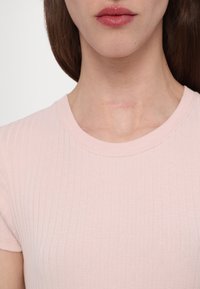 Femme portant un t-shirt côtelé rose clair, montrant une cicatrice horizontale discrète à l'avant du cou sous les lèvres et le menton.