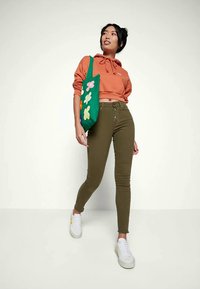 Jovem mulher vestindo um hoodie curto laranja, calças skinny de cintura alta olivas, ténis brancos e a segurar uma tote bag verde com desenhos coloridos de flores.