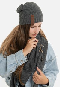 Donkergrijze gebreide beanie met een geribd ontwerp en een label. Gecombineerd met een gestructureerde grijze sjaal, voorzien van strikjes en een decoratieve kraal.