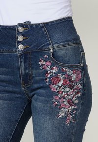 Jeans de mezclilla azul de tiro alto con bordado floral en rosa y blanco en el muslo izquierdo. Tres botones decorativos plateados en la cintura.