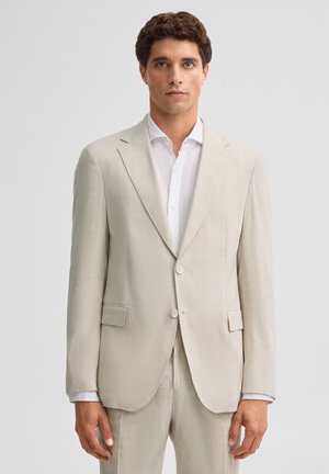 Mann trägt eine beige Anzugjacke und passende Hose über einem weißen Hemd und steht vor einem schlichten hellgrauen Hintergrund.