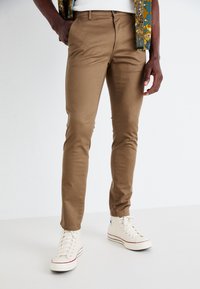 Chino étroit brun en mélange de coton, avec une texture lisse et des poches latérales, associée à des baskets montantes blanches.