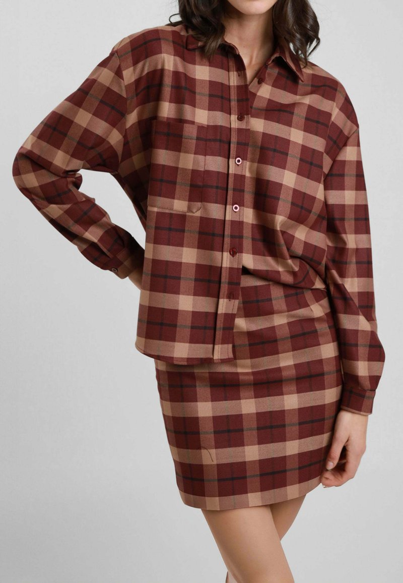 Donna che indossa una camicia a quadri rossa e beige abbinata a una gonna corta, in piedi con una mano sui fianchi contro uno sfondo semplice.