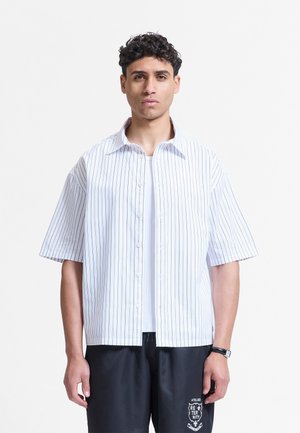 Mann mit kurzen lockigen Haaren trägt ein weißes, kurzärmeliges, gestreiftes Hemd über einem weißen T-Shirt und schwarze Hosen mit aufgedrucktem Logo.
