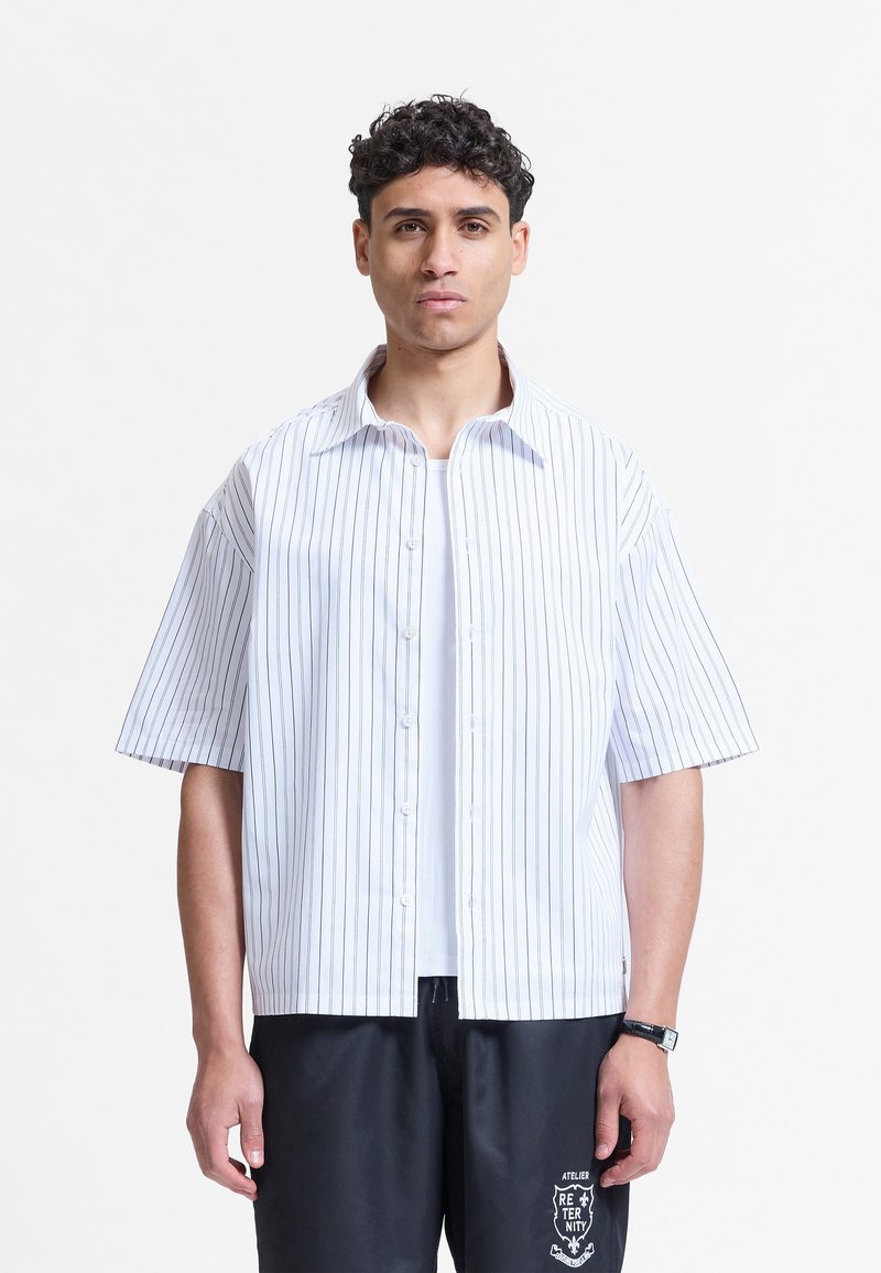 Homme aux cheveux courts et bouclés portant une chemise blanche à manches courtes rayée sur un t-shirt blanc et un pantalon noir avec un logo imprimé.
