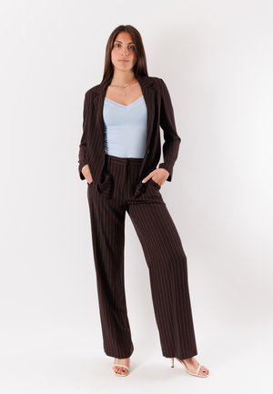 Donna in piedi con le mani nelle tasche, indossa un completo scuro a righe sottili con pantaloni larghi, top azzurro e sandali beige con tacco alto su sfondo bianco.