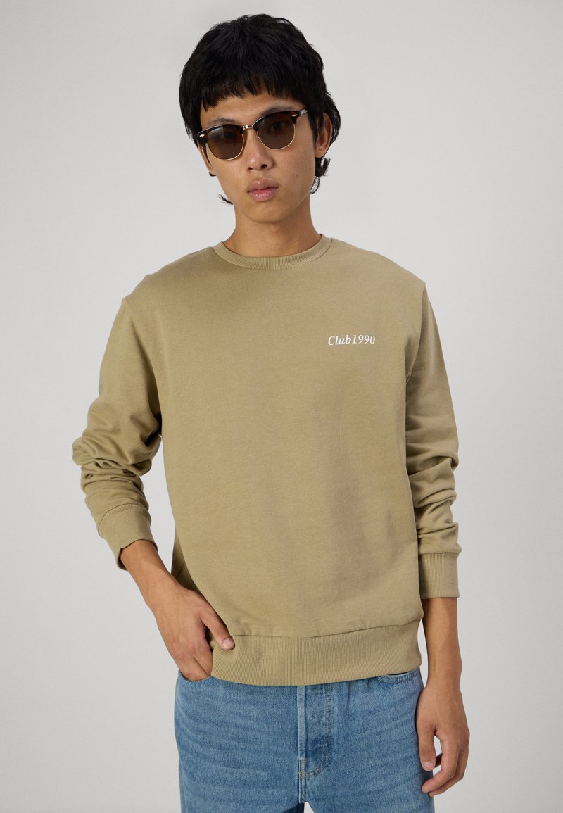 Felpa crewneck marrone chiaro con il testo bianco "Club1990"; polsini e bordo a costine; abbinata a jeans azzurri. Vestibilità casual e texture morbida.