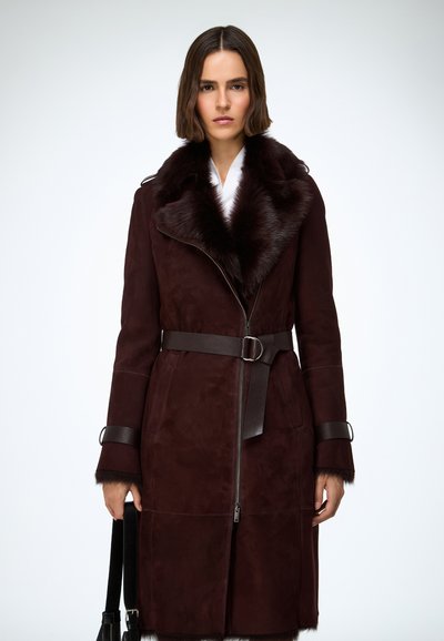 Zamatový kabát bordovej farby s jemným kožušinovým golierom, opaskom v páse a na zips vpredu. Má ozdobné manžety a stredne dlhý, štruktúrovaný strih.