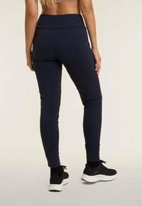 Leggings aderenti blu navy con una cintura larga, texture morbida e gambe tapered. Indossati con scarpe da ginnastica nere per creare contrasto.
