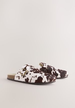 Klompe smeđe i bijele boje s uzorkom kravlje kože i kopčom na remenu. Tekstura nalik antilopu, okrugli vrh i ravni potplat. Dizajnirane za ležerno nošenje.