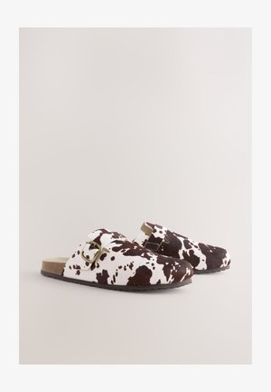 Klompe smeđe i bijele boje s uzorkom kravlje kože i kopčom na remenu. Tekstura nalik antilopu, okrugli vrh i ravni potplat. Dizajnirane za ležerno nošenje.