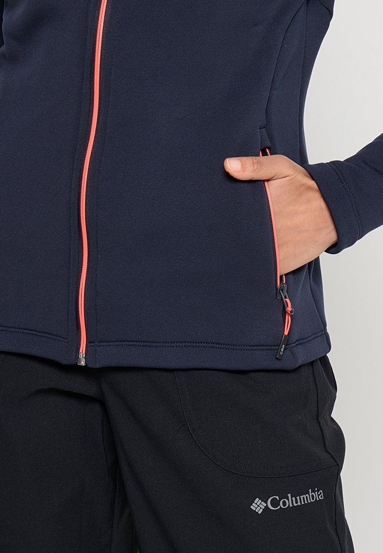 Veste zippée marine avec une fermeture éclair corail et des poches, fabriquée dans un matériau lisse. Pantalons noirs avec un logo Columbia sur le côté.