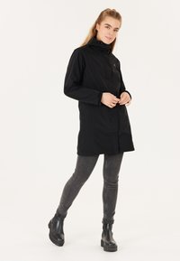 Svart vattentålig jacka med hög krage, framknappar, sidofickor och en något figursydd silhuett. Bärs över gråa skinny jeans.