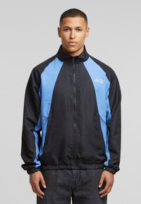 Sort og lyseblå zip-up jakke med høj krave, elastiske cuffs og en søm. Har et logo på brystet og sidelommer.