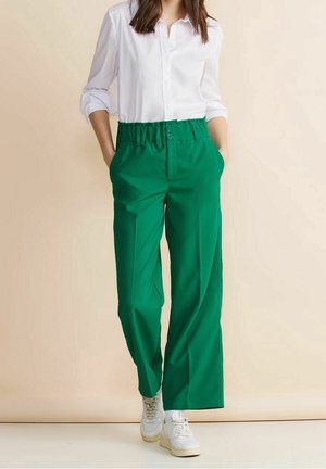 Vrouw in een wit overhemd met opgerolde mouwen, groene wijde broek met hoge taille en witte sneakers, staand met handen in de zakken.