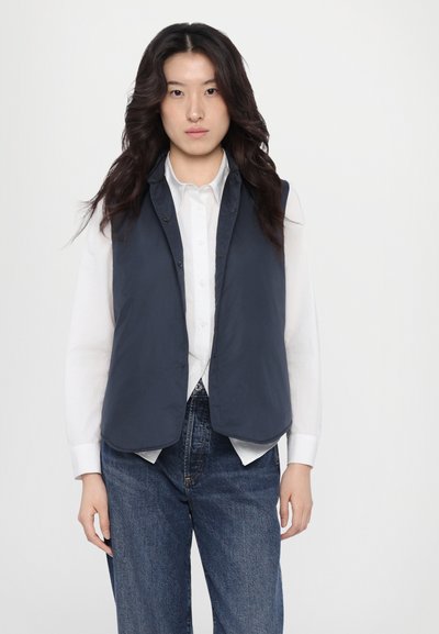 Marineblauwe mouwloze vest gemaakt van lichtgewicht materiaal, met een drukknoopsluiting, gedragen over een witte blouse en gecombineerd met donkerblauwe jeans.