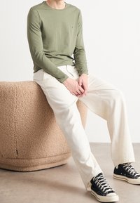 Koszula w oliwkowy kolor z długimi rękawami, białe spodnie, oraz czarne sneakersy typu high-top. Siedzi na beżowym teksturowanym pufie w neutralnym otoczeniu.