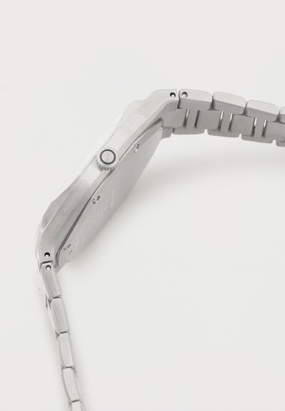 Ασημένιο μεταλλικό ρολόι χειρός, απεικονίζεται από το πλάι, δείχνοντας το στόλισμα ρολογιού και το συνδεδεμένο βραχιόλι σε λευκό φόντο.