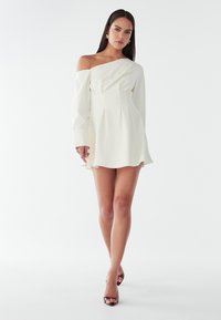 Abito mini crema con spalle scoperte, maniche lunghe, tessuto drappeggiato, vita plissettata e orlo svasato. Indossato con tacchi neri con cinturini.