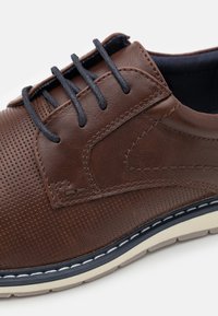 Brown leather shoe featuring perforated detailing, blue laces, and a contrasting white sole with stitching accents. Smooth texture overall.