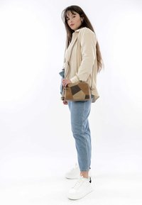 Sande- und braunfarbene geometrisch gemusterte Leder-Umhängetasche mit verstellbarem Riemen, gehalten von einer Person, die ein Creme-Oberteil und helle blaue Jeans trägt.