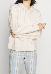 Sweat à capuche beige avec cordon de serrage, coupe décontractée, texture douce et poignets côtelés, associé à un pantalon à carreaux bleu clair.