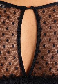 Black sheer mesh top with black dot patterns, featuring a low V-shaped back and decorative ruffle trim along the hem.