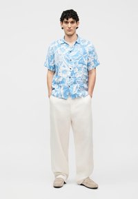 Mann steht mit in den Taschen steckenden Händen, trägt ein blaues kurzärmeliges Hemd mit Blumenmuster, lockere weiße Hosen und beige Schuhe vor weißem Hintergrund.