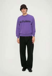 Purple sweatshirt with "No Problemo" in black lettering, ribbed cuffs and hem, paired with black trousers and black shoes.