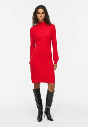 Femme aux cheveux bouclés portant une robe rouge moulante à manches longues et des bottes noires montantes à talons, debout devant un fond blanc.